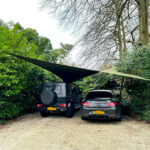 Two cars parked on a gravel driveway beneath a large triangular shade sail stretched between tall posts, surrounded by dense green hedges and trees.
