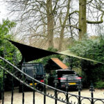Black SUV and a blue SUV parked on a gravel driveway under a tan shade sail, with a wrought-iron gate and lush hedges in a garden setting.