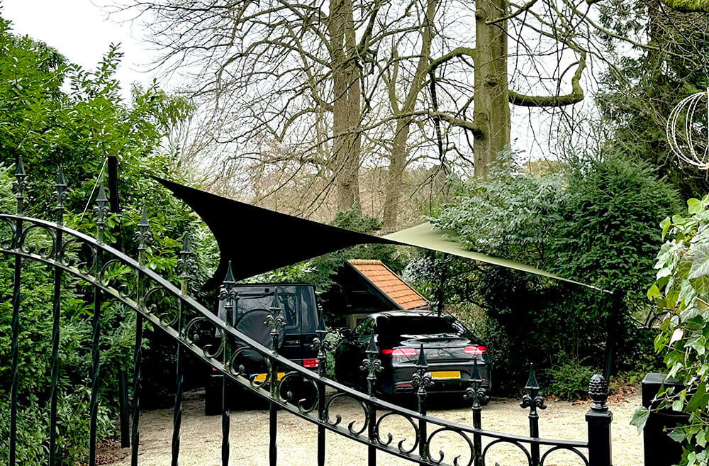 Black SUV and a blue SUV parked on a gravel driveway under a tan shade sail, with a wrought-iron gate and lush hedges in a garden setting.