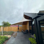 Modern backyard patio with a white fabric shade sail, dark seating area, and wooden fence beside a glass door entrance.