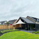 Modern house with a white fabric shade sail over a patio seating area and a manicured lawn in the foreground.