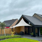 Modern house with a white fabric shade sail over a patio and black outdoor furniture on a gray stone patio. Lush green lawn in foreground and wooden fence.