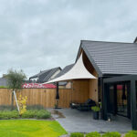 Backyard patio with a white sail shade, wooden fence, and outdoor seating on gray stone tiles beside a modern house.