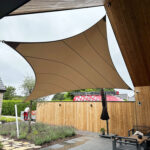 Large beige fabric shade sail stretched above a patio area with wooden walls and outdoor seating.