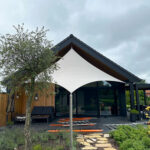 Modern house with a white triangular shade sail over the patio; orange ladders laid on the ground and a stone stepping path leading to the entrance.