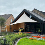 Backyard of a modern house with a white tensile shade sail over the patio, green lawn, and construction frames on the ground nearby.