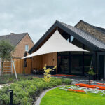 Modern house with a black-framed glass patio and a white triangular shade sail over the outdoor seating area, lawn and garden beds nearby.