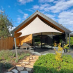 Modern house with a large beige shade sail over a patio and seating area, under a blue sky with clouds.