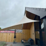 Patio seating area under white fabric shade sails attached to a wooden house exterior.