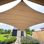 Tan fabric shade sail stretched under a wooden overhang, shielding a stone pathway in a landscaped garden area.