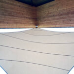 Corner of a wooden pavilion interior with large white fabric shade sails creating curved panels overhead.