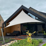 White tensile shade sail spanning a glass-front patio, attached to tall posts over outdoor seating near a modern house; stone path and landscaping in foreground.