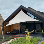 White triangular shade sail over a modern wooden house patio with outdoor seating and a garden path.