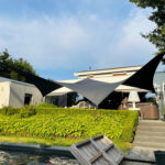 Backyard patio under black-and-white geometric shade sails over seating area, with hedges and steps to the house; a water feature is in the foreground.