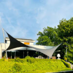 Modern house with two large fabric shade sails over a patio, beside a water feature and trimmed hedges.