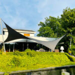 Modern house with a patio shaded by black and white fabric sails over a hedge beside a water feature.
