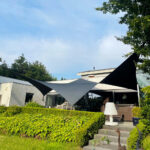 Modern single-story home with black and gray triangular shade sails over a patio area, hedges in front.