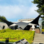 Outdoor patio with black-and-white sail shade over a seating area, hedge in the foreground, and a modern house behind; sunny day with blue sky.