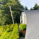 Path along a building with a large gray shade sail, green hedges, and potted plants.