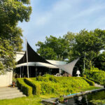 Outdoor cafe area with black and white shade sails over seating, hedges, and a pond with water lilies in the foreground.