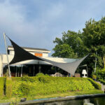 Modern house with black-and-white fabric shade sails over an outdoor seating area, next to a trimmed hedge and pond.