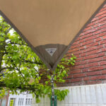 Beige outdoor shade sail attached to a brick wall with green leaves nearby and a street view beyond.