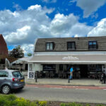 Cafe with outdoor seating under a beige awning on a sunny street, a silver car parked in front and a blue sky with clouds above.