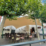 Outdoor cafe seating under a large beige canopy with green trees and light brick walls in the background.
