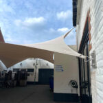 Outdoor courtyard with beige fabric shade sails attached to white brick buildings; stacked chairs visible on the left side.