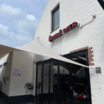 Exterior view of a white brick building with a red-and-black 'Brand Bier' sign above a glass entrance under a large white canopy