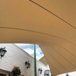 Beige curved canopy over a white brick courtyard with hanging flower basket and black lanterns along the wall.