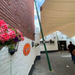 White brick building with an orange sign reading 'CINEY', pink flowers, and a large beige canopy supported by green poles in an outdoor alley.