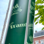 Close-up of a green advertising banner wrapped around a pole, showing 'Café Tram...' and beer logos with leaves in the background; a rope is on the left side.