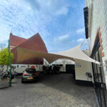 White fabric shade sail spans a courtyard parking area between white brick walls and a red-tiled roof building, with a blue sky above.
