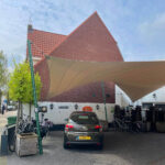Car parked under a large beige sail shade in front of a red brick building with a steep gabled roof, bicycles nearby, and a white wall with lamps in the background.