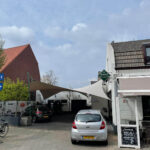 Silver hatchback parked on a cobblestone street beside a white storefront with a green diamond sign and beige shade sail overhead.