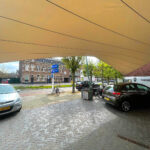 Curved tan fabric canopy arching over a cobblestone street with parked cars and bicycles, brick buildings in the background.