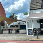 Outdoor cafe seating area with beige sails, planters of flowers, and a chalkboard menu outside a white storefront called Brand on a sunny street.