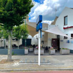 Outdoor cafe seating along a white brick building with blue signpost and 'Brand Bier' sign, green trees, and planters under a shade sail.