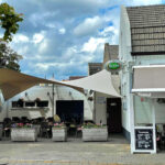 Outdoor cafe with beige shade sails, white brick buildings, and flower planters; diners sit at tables under the awning.