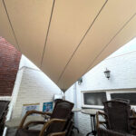 Outdoor cafe seating under a large beige canopy over white brick walls, with wicker chairs and round tables.