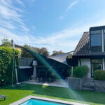 Backyard with a bright blue rectangular pool, green lawn, and a modern house with a tiled roof on the right under a clear blue sky.