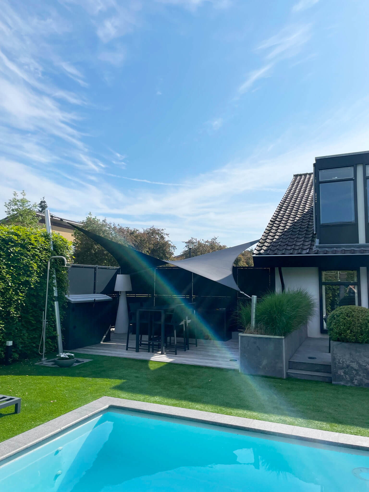 Drutenkey-doek-luxe-zeildoek-terras-overkapping-op-maat-stormvast-verwijderbaar-01 Backyard with a bright blue rectangular pool, green lawn, and a modern house with a tiled roof on the right under a clear blue sky.