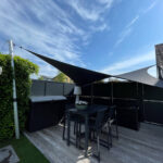 Backyard patio with a black shade sail over a high outdoor table and chairs, against a dark privacy fence and blue sky.
