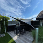 Outdoor backyard patio with black table and high chairs under white shade sails, wooden deck and tall planters, blue sky above.