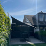Modern backyard patio with a black shade sail, wooden deck, and outdoor furniture against a bright blue sky.