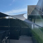 Patio scene with dark privacy fence, wooden deck, and outdoor dining set beneath curved fabric shade against a blue sky.