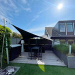 Backyard patio with black shade sails over a dining table set on a wooden deck, modern house with large windows in the background, under a bright blue sky.