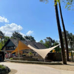Outdoor plaza with a pitched-roof building and beige triangular shade sails under a bright blue sky.