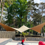 Outdoor courtyard with beige triangular shade sails stretched over a stone-paved area and a low stone wall, surrounded by tall trees.